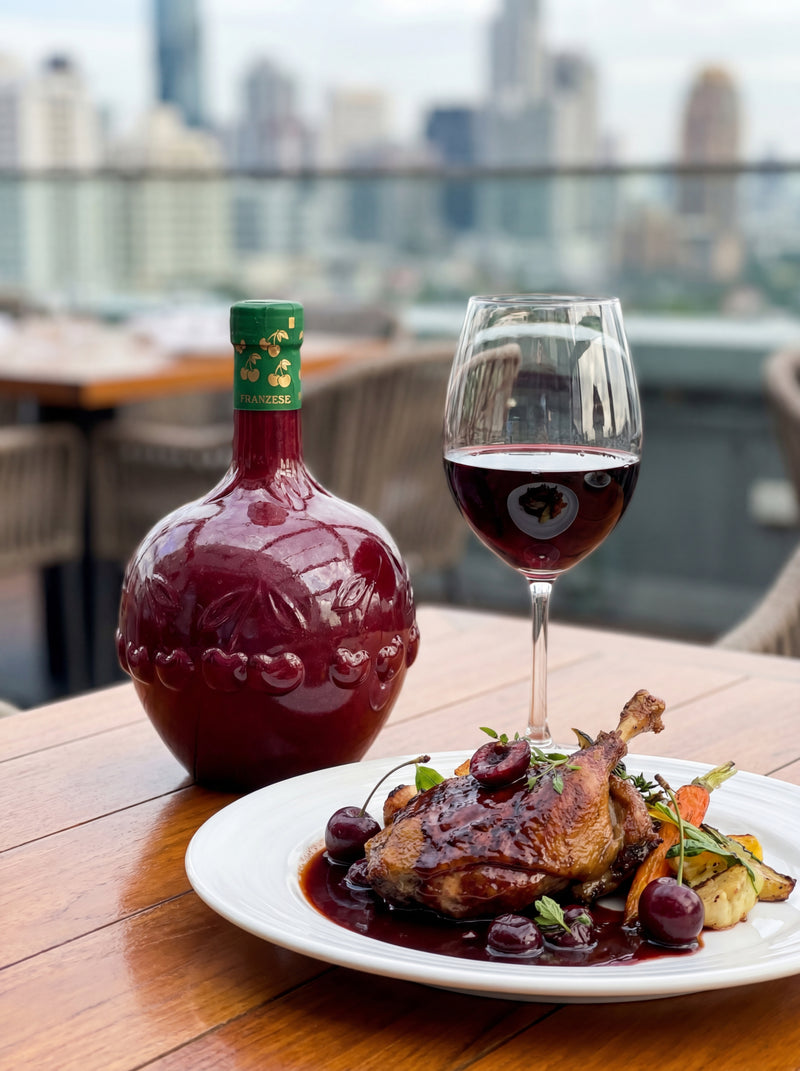 Dinner setting with a bottle of cherry wine, a glass of cherry wine, and a dish of roasted chicken with vegetables on a table with a cityscape background.