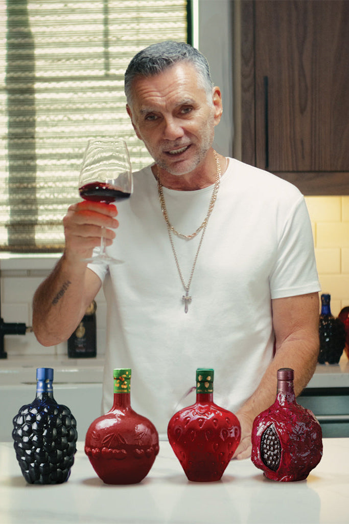Man holding a glass of red wine in a kitchen with four bottles on the counter.