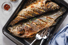 Two grilled fish on a baking tray with seasonings and utensils.