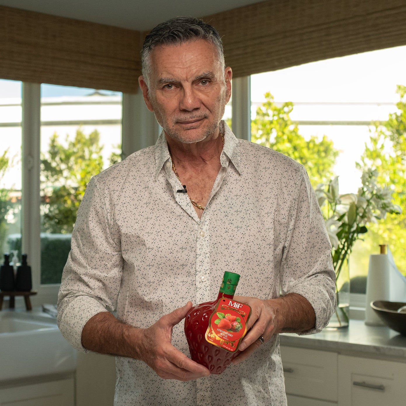 Man holding a red bottle in a kitchen with large windows showing greenery outside.